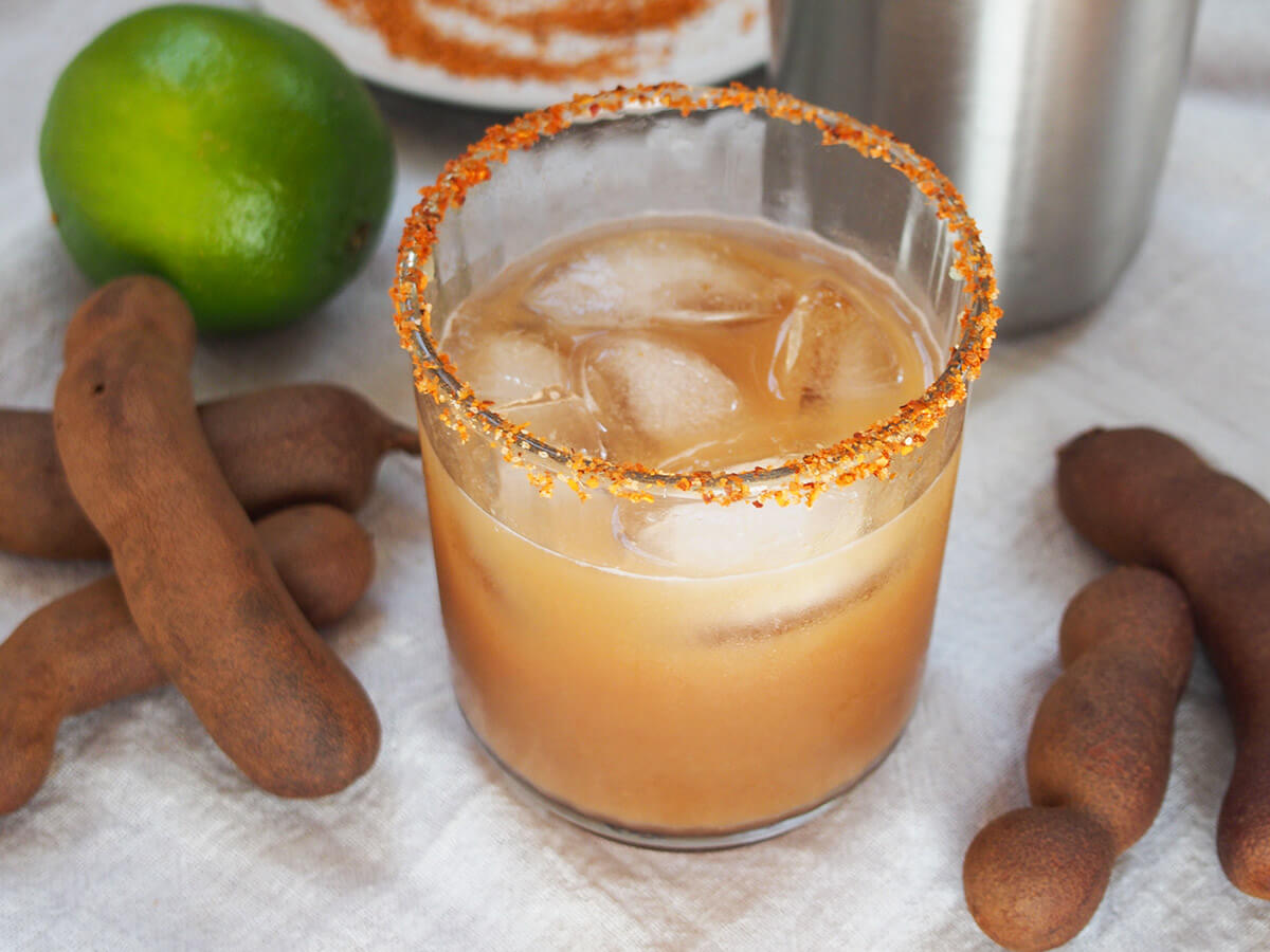This tamarind margarita gets an extra burst of sweet tart flavor from citrus-like tamarind, that also adds depth to the drink. It's no wonder this is a favorite variation on the classic cocktail. Perfect icy-cold on a warm day. glass of tamarind margarita with tamarind pods either side of glass and lime behind