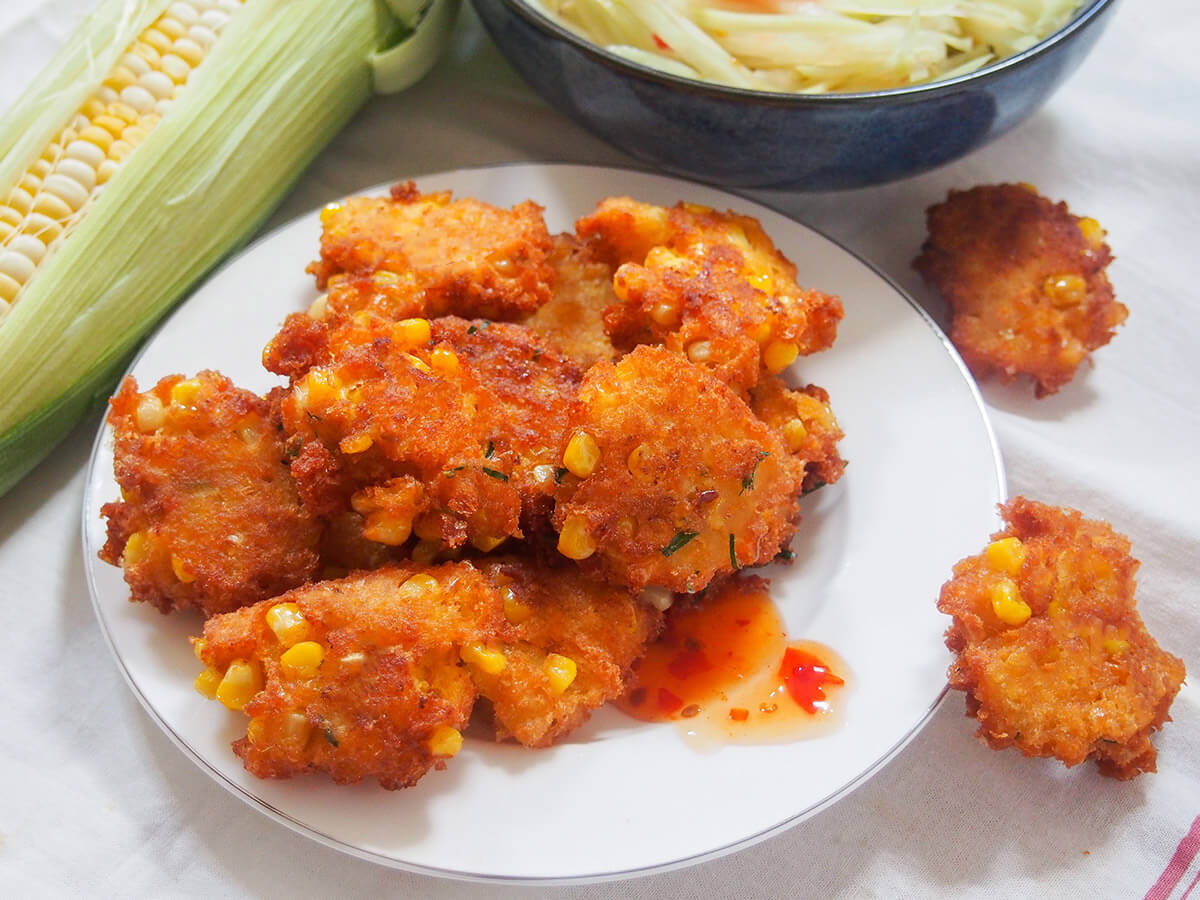 These Thai corn fritters (tod man khao pod) are addictively tasty little bites, packed with corn and flavored with bright lime leaves and red curry paste. They're easy to make, naturally gluten free and perfect for snacking on, or as an appetizer. plate of Thai corn fritters with cob of corn to side