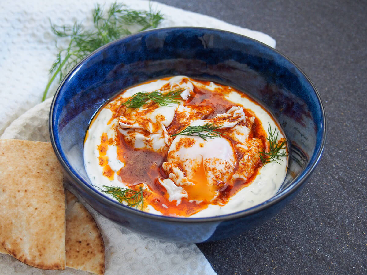 These Turkish eggs, çilbir, are so easy to prepare and have a wonderful combination of flavors. Poached eggs are served over a garlicy yogurt base, topped with a warm-spiced butter. It's light and thoroughly delicious. bowl of Turkish eggs cilbir with yolk of front egg leaking into sauce