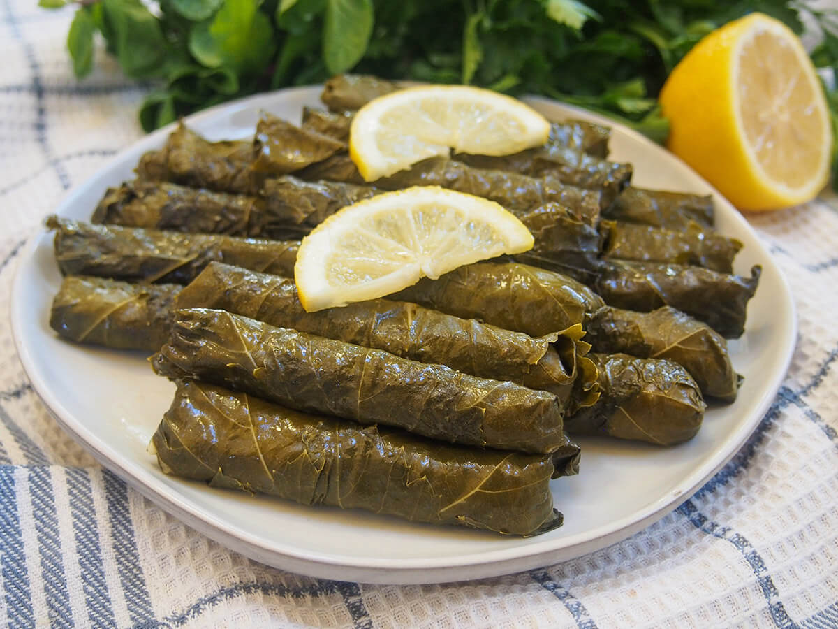 These Turkish stuffed grape leaves, yalancı dolma, have a simple vegetarian filling of mainly rice, herbs and currants. They're bright, tasty and comforting - a great addition to any meze platter. plate of Turkish stuffed grape leaves, yalanci dolma with lemon and herbs behind