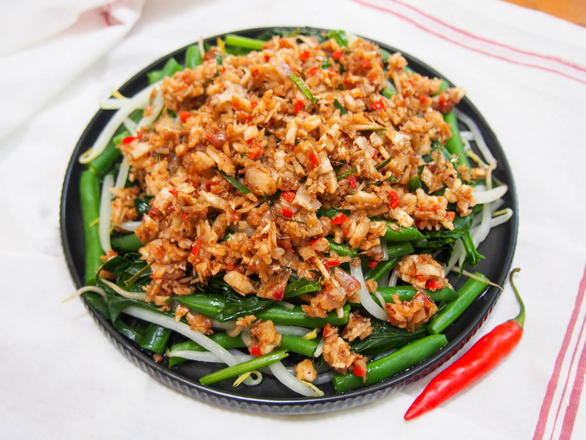 Urap sayur is a popular Indonesian dish combining blanched vegetables with a spiced coconut topping. It's easy to make with a delicious combination of spicy, sweet and sour flavors. plate of Urap sayur (Indonesian vegetables with coconut topping) with red chili to side