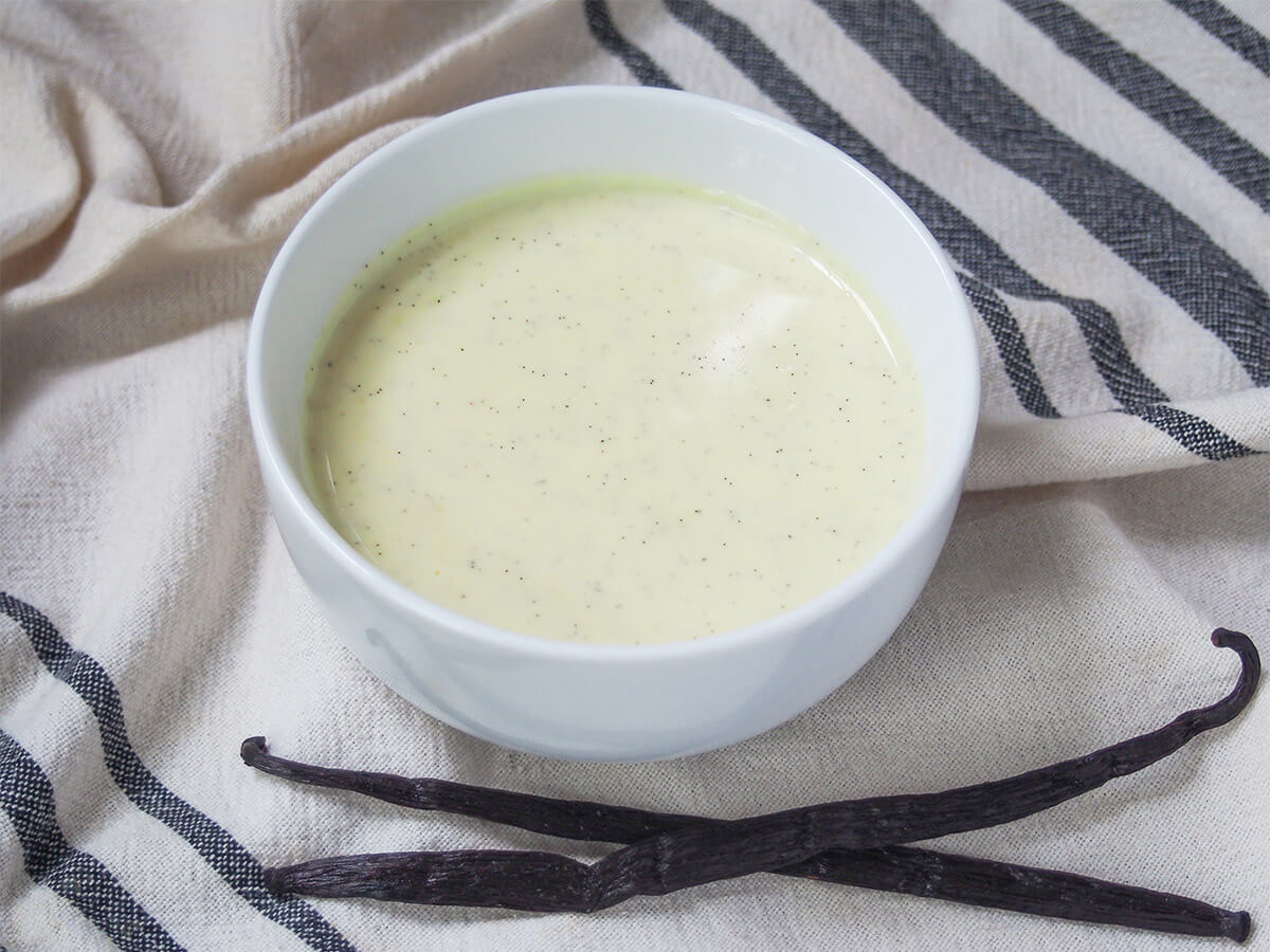 Vanillesoβe, German vanilla sauce, is easy to make and great served warm or chilled, poured over all your favorite desserts and treats. Creamy, comforting and delicious. bowl of Vanillesosse German vanilla sauce with two whole vanilla pods below