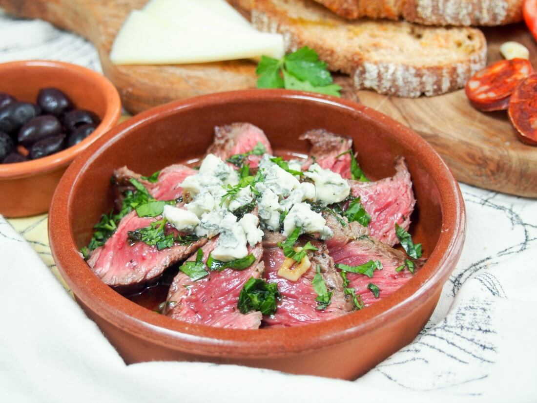 This white wine marinated steak with blue cheese is easy to make, full of flavor and makes a great tapas dish alongside your favorites or make it a main. dish with white wine marinated steak topped with blue cheese with some olives and bread behind