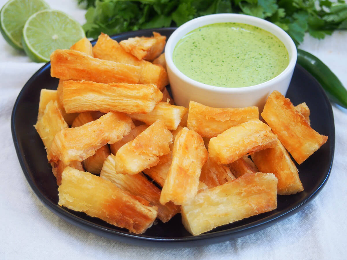 Yuca fries are wonderfully crisp on the outside and gently soft in the middle. They are easy to make and just begging to be dipped in your favorite sauce. plate of yuca fries with bowl of Peruvian green sauce at the back and cut lime behind plate